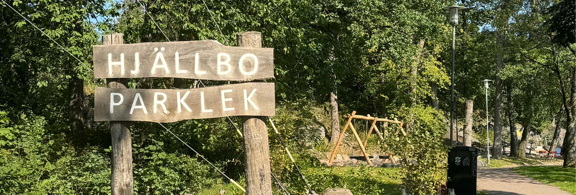 En lekpark omgiven av lummiga trä. I förgrunden står en rustik träskylt som säger "Parklek Hjällbo".